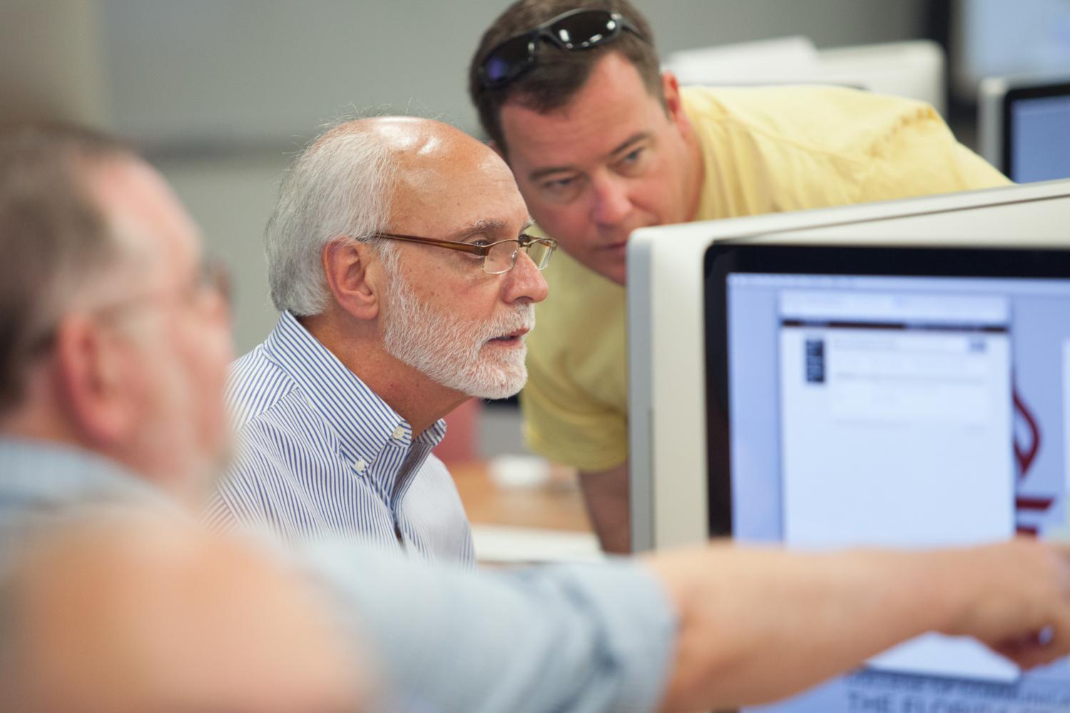 "Three older men seated at a table, scrutinizing information on computer monitors"