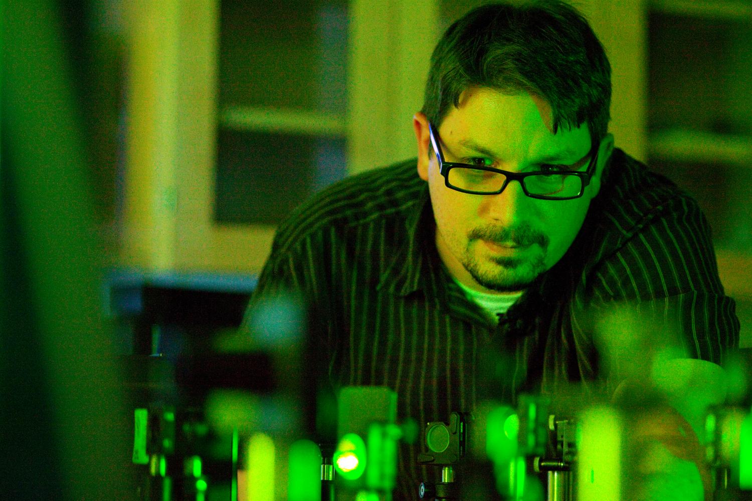A male scientist wearing black glasses examines green, fiber optic lasers.