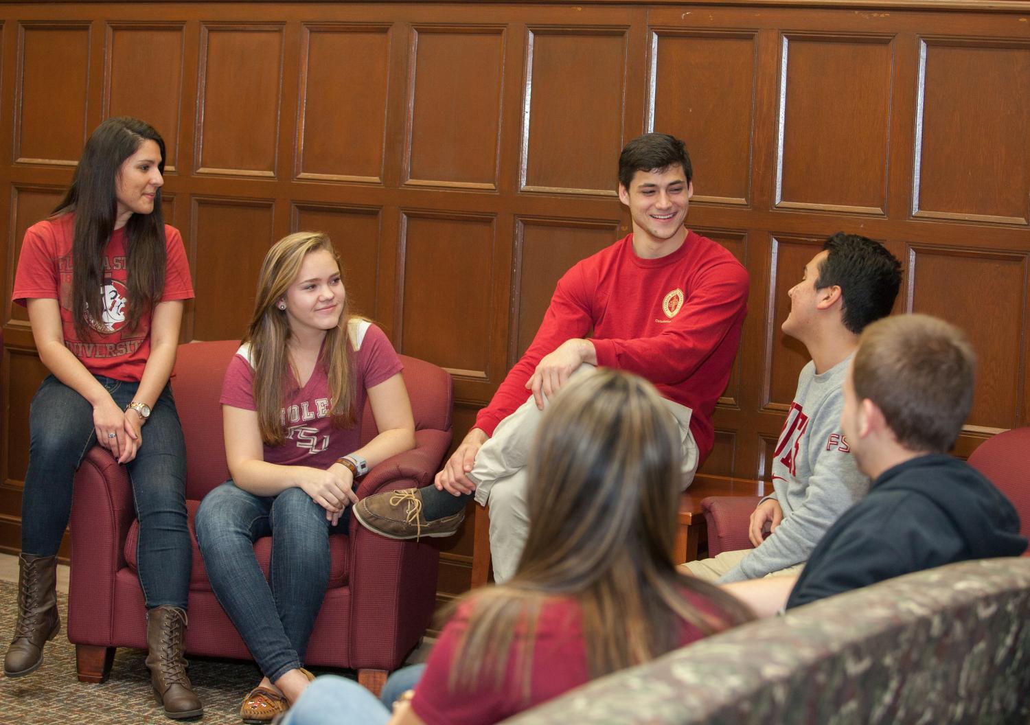 "A group of six students sitting on garnet armchairs smiling and talking amongst themselves"
