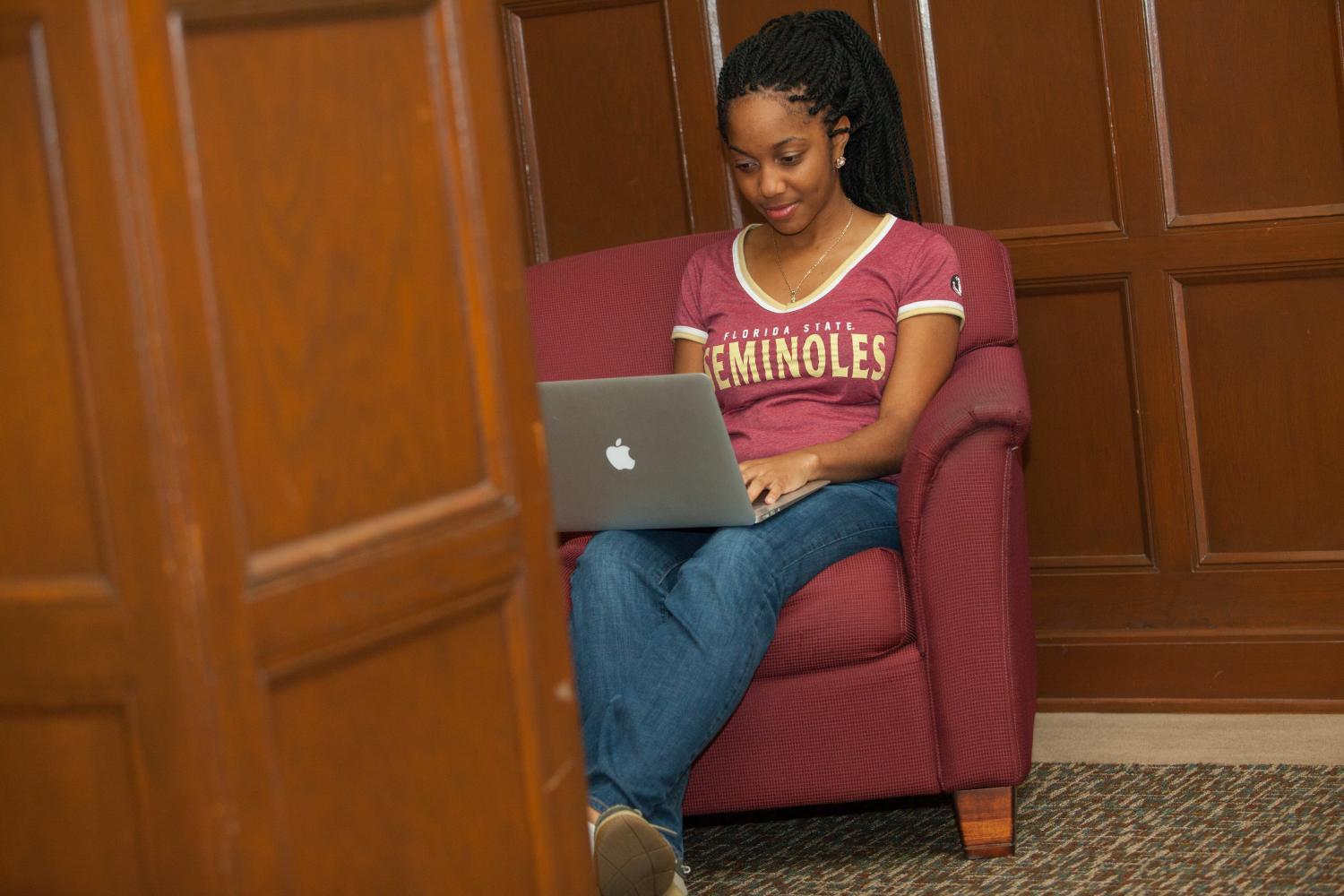A student smiling at her MacBook while seated in a comfortable garnet chair.