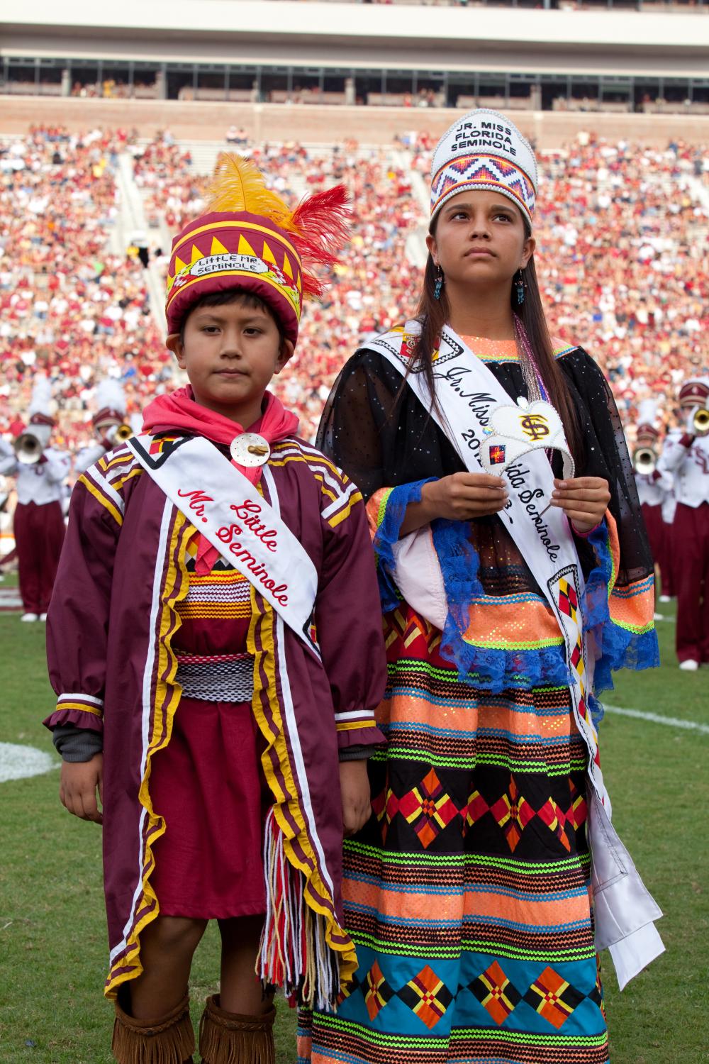 Little Mr. Seminole and Jr. Miss Florida Seminole stand together in the middle of Doak Campbell stadium's field. They are wearing traditional regalia under their pageant sashes.
