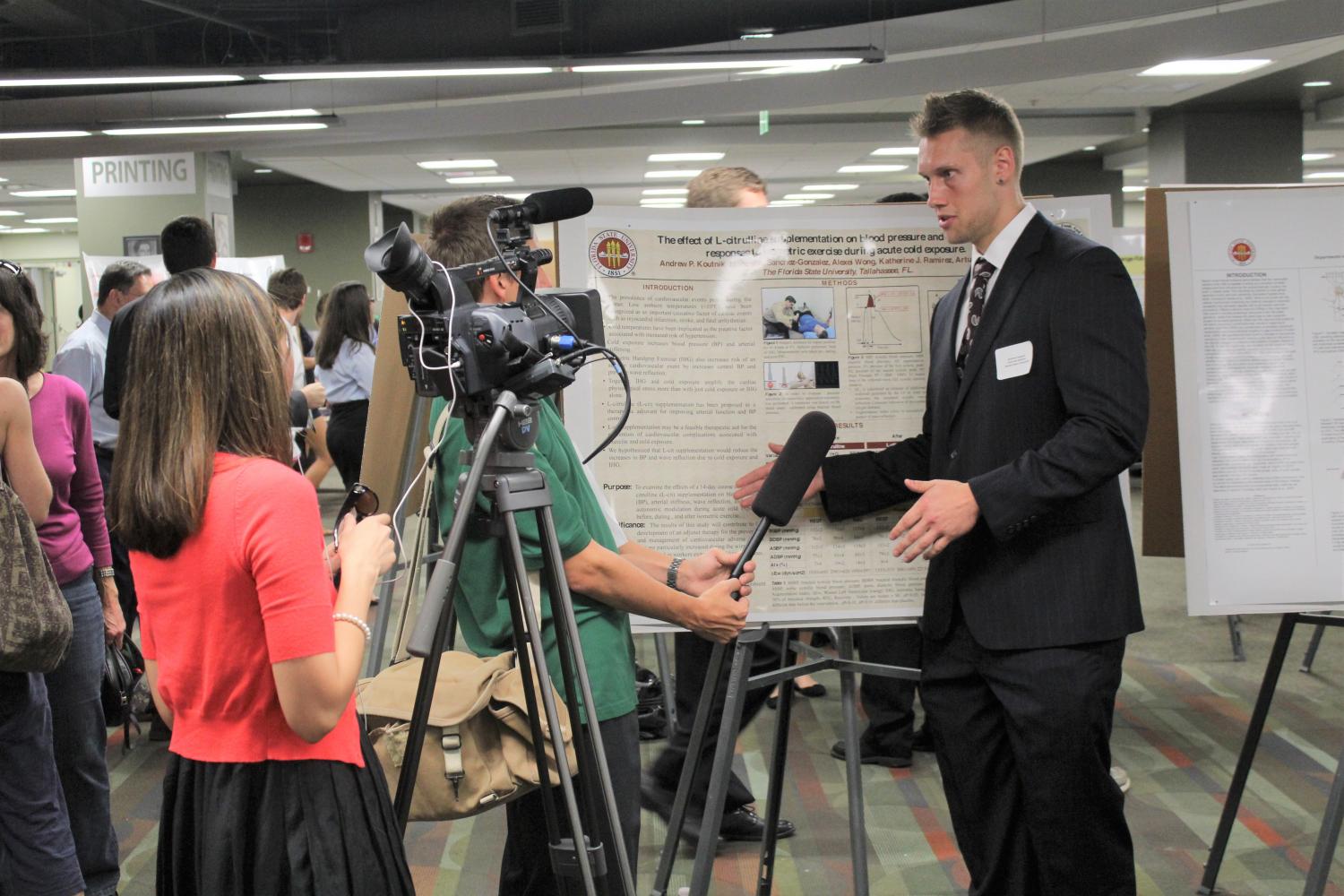 A student wearing a black suit is interviewed about his research presentation by a news crew. One person holds the microphone while another controls a camcorder. The student is surrounded by event guests.