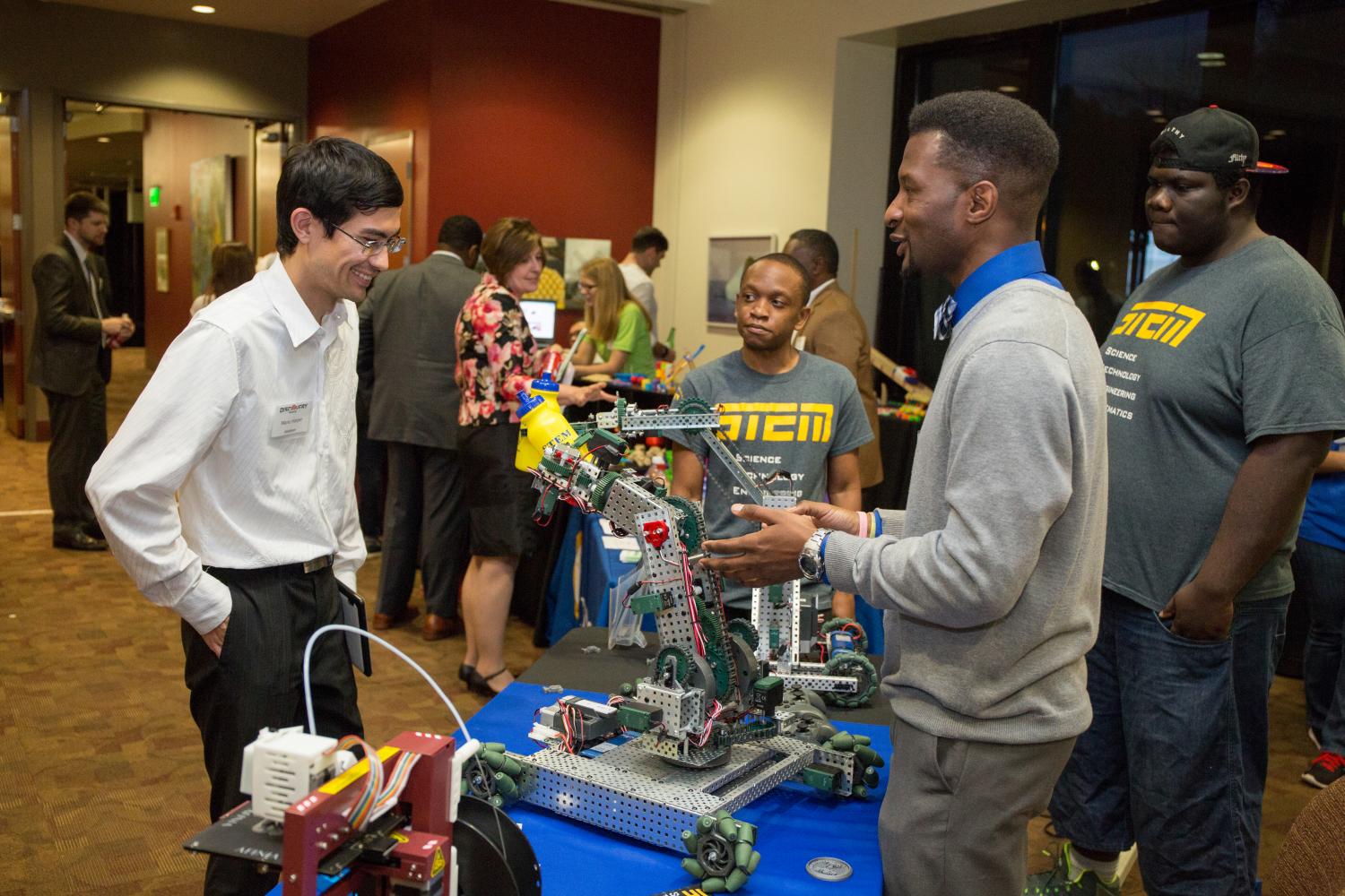 Three students excitedly present their robot project to an exhibitor at a conference.